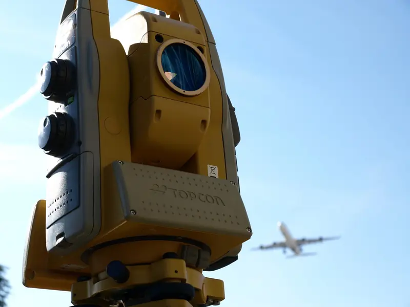 Station totale Topcon jaune sur trépied devant un ciel bleu, un avion en arrière-plan
